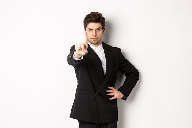 Portret van een serieuze knappe man in een pak, die één vinger laat zien om iets te verbieden of af te wijzen, te zeggen dat hij moet stoppen, het niet met je eens is, op een witte achtergrond staat.