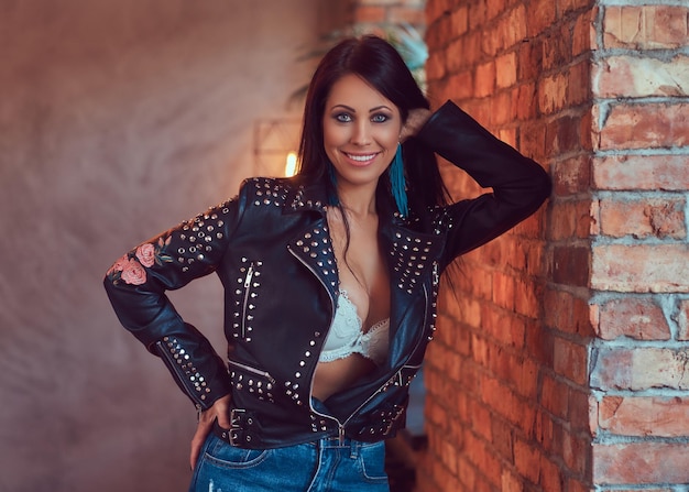 Portret van een sexy sensuele brunette poseren in stijlvolle leren jas en jeans leunend tegen een bakstenen muur.