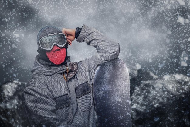 Portret van een snowboarder gekleed in een volledig beschermende uitrusting voor extreem snowboarden, poserend met snowboard tegen de achtergrond van bergen