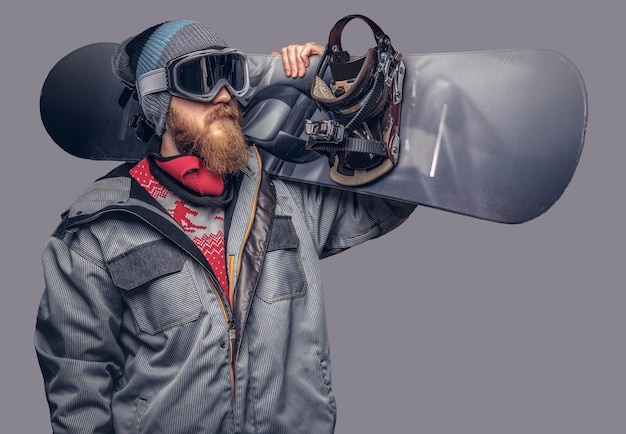 Portret van een snowboarder gekleed in een volledige beschermende uitrusting voor extreme snowboarden poseren met een snowboard op zijn schouder in een studio. Geïsoleerd op een grijze achtergrond.