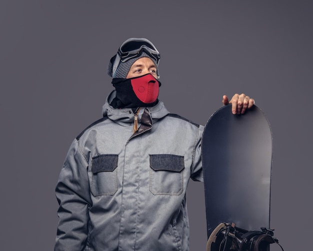 Portret van een snowboarder gekleed in een volledige beschermende uitrusting voor extreme snowboarden poseren met snowboard in een studio. Geïsoleerd op een grijze achtergrond.