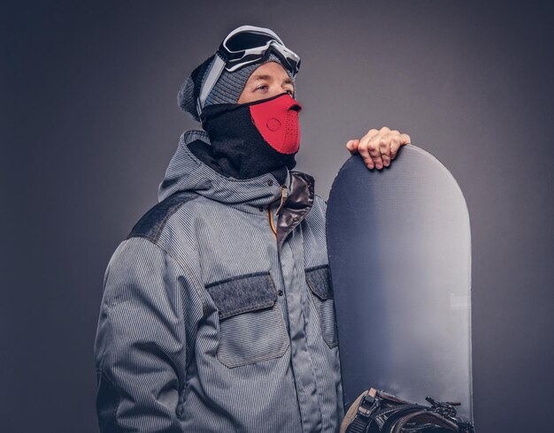 Portret van een snowboarder gekleed in een volledige beschermende uitrusting voor extreme snowboarden poseren met snowboard in een studio. Geïsoleerd op een grijze achtergrond.