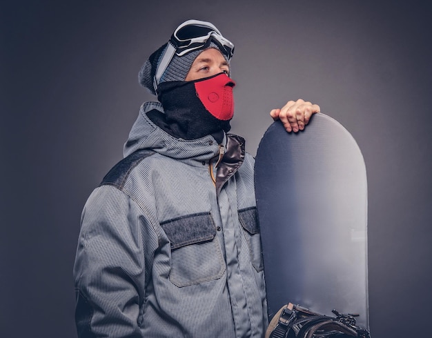 Portret van een snowboarder gekleed in een volledige beschermende uitrusting voor extreme snowboarden poseren met snowboard in een studio. Geïsoleerd op een grijze achtergrond.