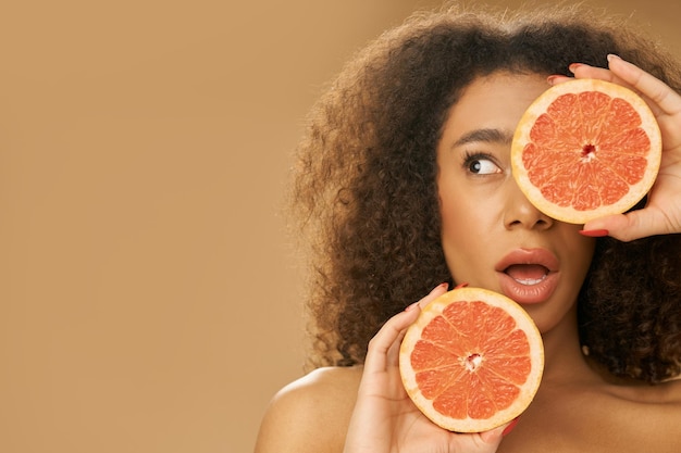 Portret van een speelse jonge vrouw van gemengd ras die verbaasd kijkt en één oog bedekt met een grapefruit-cut