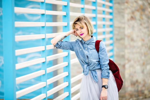 Portret van een staande glimlachende vrouw met kort blond haar, felroze lippen en naakt make-up leunend op blauwe en witte strepen hek