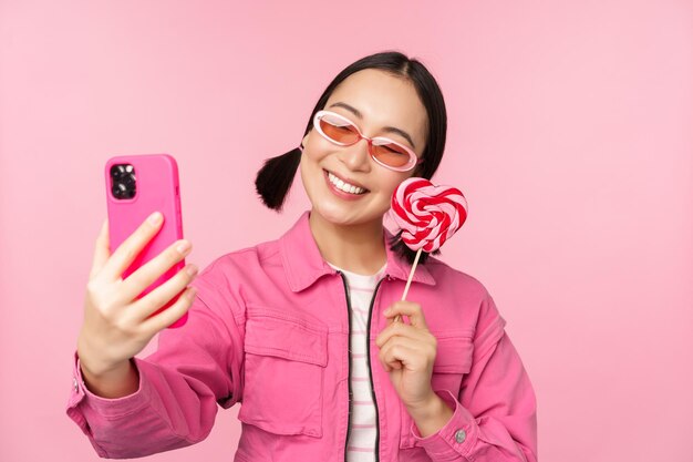 Portret van een stijlvol, gelukkig Aziatisch meisje dat selfie neemt met snoep lolipops en glimlachend foto's neemt met een mobiele app die over een roze achtergrond staat