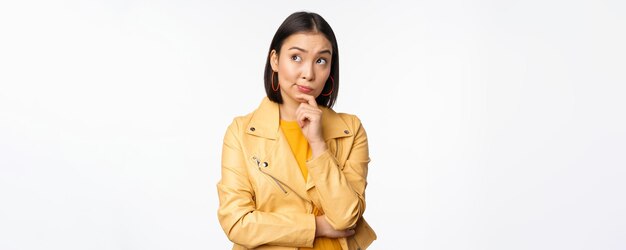 Portret van een stijlvolle Koreaanse vrouw in een gele jas die doordacht nadenkt en omhoog kijkt naar een logo of advertentie die op een witte achtergrond staat