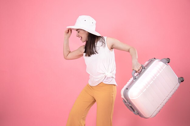 portret van een stijlvolle vrouw in modieuze zomerkleding en een witte hoed op roze met een koffer om te reizen.