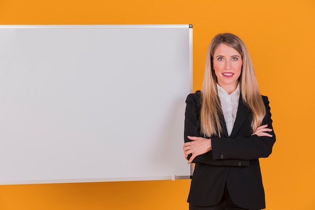 Gratis foto portret van een succesvolle jonge onderneemster die zich dichtbij whiteboard tegen een oranje achtergrond bevindt