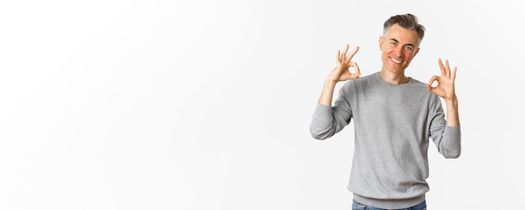 Gratis Foto portret van een succesvolle knappe man in een grijze trui die goede tekenen vertoont en tevreden glimlacht, keurt iets goeds goed dat een product aanbeveelt dat over een witte achtergrond staat