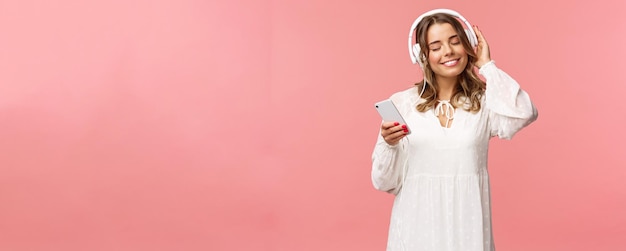 Portret van een teder gelukkig en schattig blond meisje in een witte jurk sluit de ogen en glimlacht tevreden terwijl je naar muziek luistert in een koptelefoon die warmte en geluk voelt en geniet van favoriete track hold mobiele telefoon