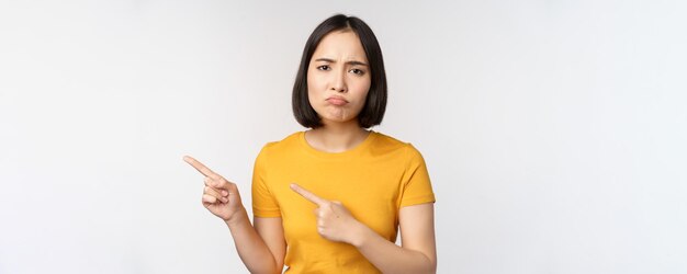 Portret van een teleurgesteld brunette aziatisch meisje dat met de vingers wijst en grimassen van streek raakt en iets onaangenaams over een witte achtergrond laat zien