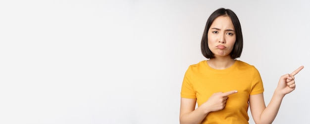Portret van een teleurgesteld, humeurig Aziatisch meisje dat met de vingers naar rechts wijst en naar een oneerlijke klacht kijkt