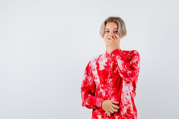 Portret van een tienerblonde man die de mond bedekt met de hand in een te groot hemd en er een gelukzalig vooraanzicht uitziet