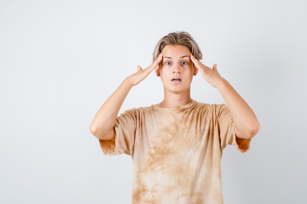 Portret van een tienerjongen die de handen op de tempels in een t-shirt houdt en een verbaasd vooraanzicht kijkt