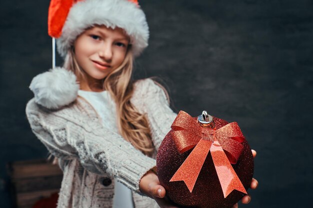 Portret van een tienerjongen met een kerstmuts met een grote kerstbal op een donkere gestructureerde achtergrond.