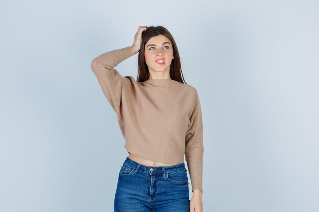 Portret van een tienermeisje dat de hand op het hoofd houdt, op de lip bijt, omhoog kijkt in trui, spijkerbroek en peinzend vooraanzicht