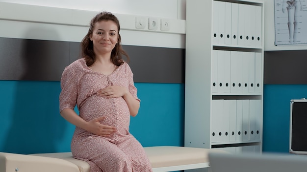 Portret van een toekomstige moeder met een babybuil die op een kastbed zit, wachtend op een consult van de dokter voor de gezondheidszorg. Zwangere vrouw die een controleafspraak op kantoor bijwoont.