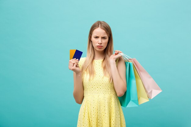 Portret van een trieste vrouw met boodschappentassen en bankkaart geïsoleerd op een blauwe achtergrond