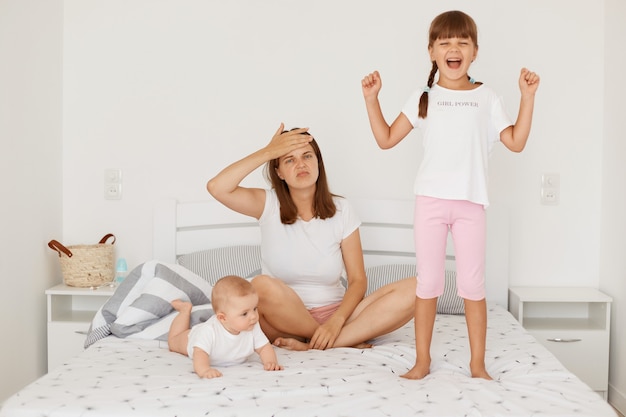Portret van een uitgeputte vrouw met een wit t-shirt op bed met haar twee luide dochters, hand op het voorhoofd, moe om veel tijd met kinderen door te brengen.