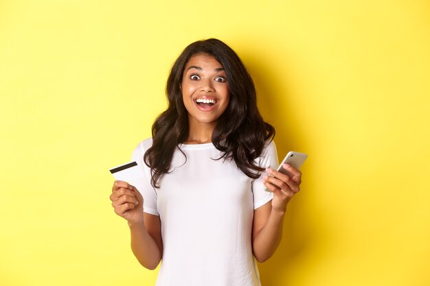 Portret van een verbaasd Afro-Amerikaans meisje met smartphone en creditcard die over gele rug staat...