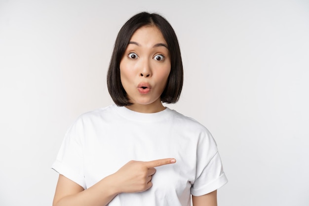Portret van een verbaasde aziatische vrouw zegt wow, wijzend naar een geweldige productaanbieding of een logo dat op een witte achtergrond staat