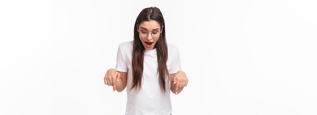 Portret van een verbaasde, nieuwsgierige, gelukkige brunette vrouw in een t-shirtbril die kijkt en geamuseerd naar beneden wijst