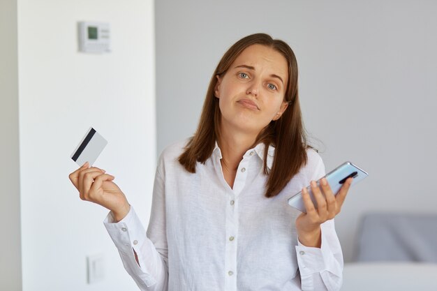 Portret van een verbaasde vrouw met donker haar die met mobiele telefoon en creditcard in handen staat, schouders ophaalt, weet niet hoe al het geld van haar bankkaart is uitgegeven.