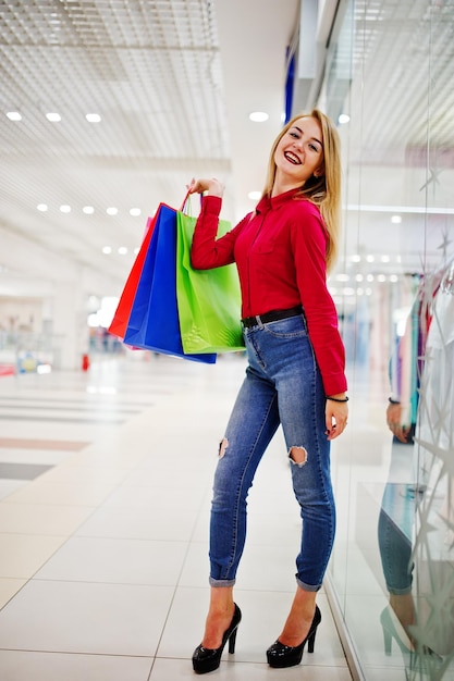 Portret van een verbluffende jonge vrouw in een rode blouse, gescheurde casual jeans en hoge hakken poseren met boodschappentassen in het winkelcentrum