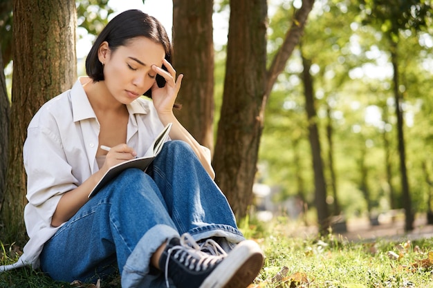 Portret van een verdrietig aziatisch meisje dat in haar dagboek schrijft en zich ongemakkelijk voelt terwijl ze alleen in het park zit onder de boom