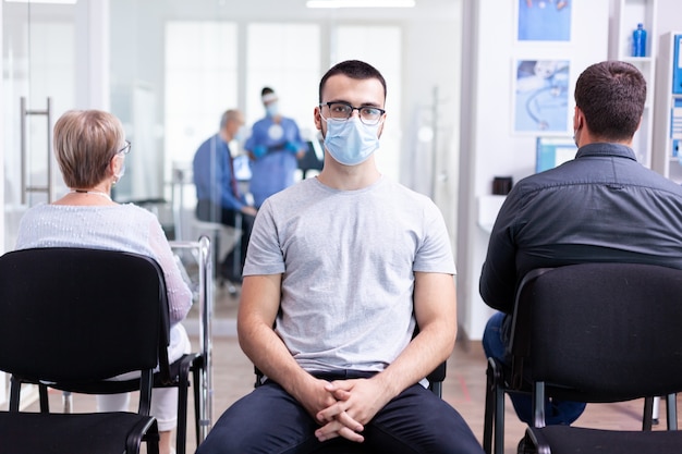 Portret van een vermoeide jongeman met gezichtsmasker tegen coronavirus in de wachtruimte van het ziekenhuis, kijkend naar de camera