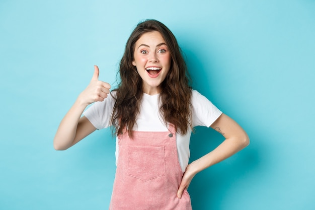 Portret van een verrast en opgewonden brunette meisje dat duim omhoog laat zien en ja zegt, gefascineerd naar adem snakkend, geweldig werk prijzend, staande tegen een blauwe achtergrond.