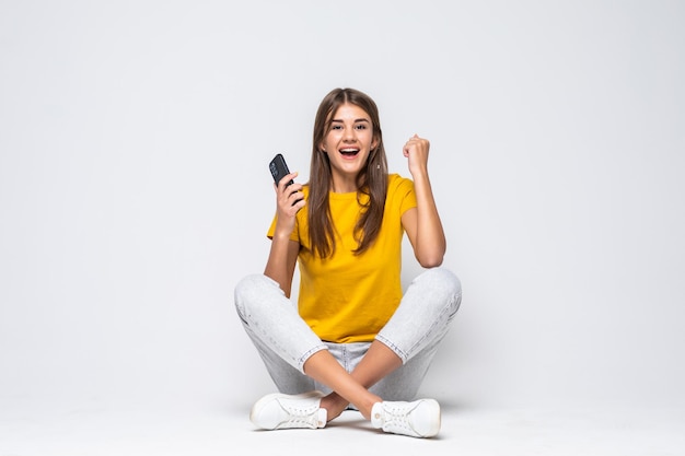 Portret van een verrast jong meisje dat telefoon gebruikt terwijl ze zit met gekruiste benen op wit wordt geïsoleerd