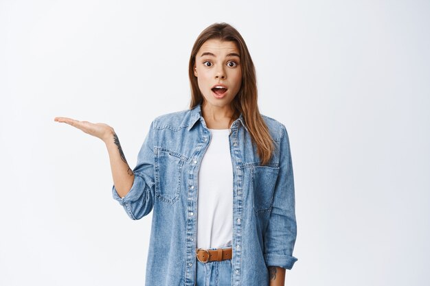 Portret van een verrast mooi meisje dat in de hand houdt, de hand zijwaarts spreidt en een object in de palm toont en verbaasd glimlacht, staande op een witte muur