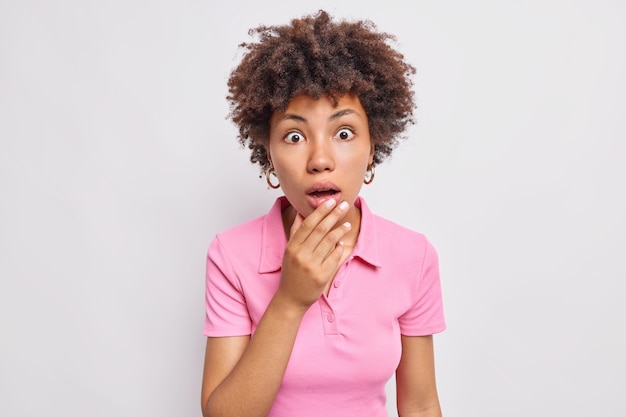 Portret van een verraste Afro-Amerikaanse vrouw hapt naar adem van verwondering en staart sprakeloos van ontzag en ongeloof kan niet geloven dat haar ogen een casual t-shirt dragen dat over een witte muur wordt geïsoleerd