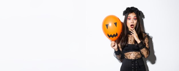 Portret van een verraste aziatische vrouw in halloween-kostuum verkleed als heks die oranje ballon vasthoudt