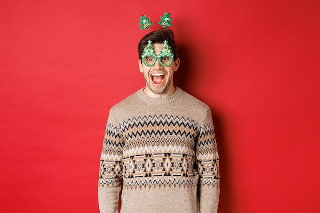 Gratis foto portret van een verraste en opgewonden knappe jongen in een feestbril en een wintertrui, die kerst viert en plezier heeft, staande tegen een rode achtergrond