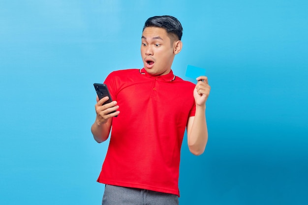 Portret van een verraste jonge aziatische man die een mobiele telefoon vasthoudt en een creditcard toont die op een blauwe achtergrond wordt geïsoleerd