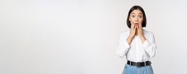 Portret van een verraste jonge kantoorvrouw, een aziatische zakenvrouw, hijgend verbaasd en zegt wow staande onder de indruk van nieuws tegen een witte achtergrond
