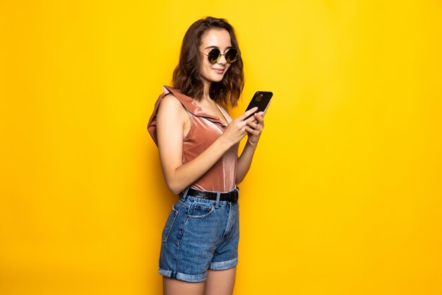 Portret van een verraste jonge vrouw in de zomerhoed en zonnebril die mobiele telefoon met behulp van die over gele muur wordt geïsoleerd