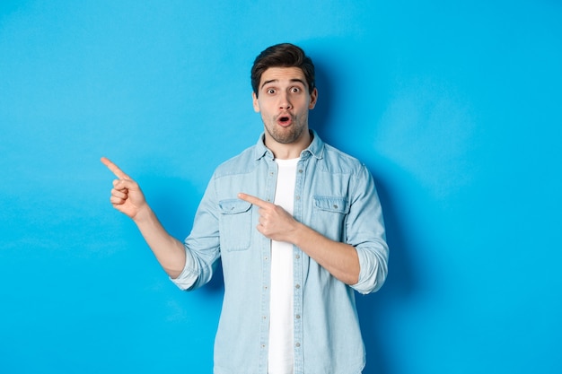 Portret van een verraste volwassen man in vrijetijdskleding die aankondiging toont, met de vingers naar links wijst en verbaasd kijkt, staande tegen een blauwe achtergrond