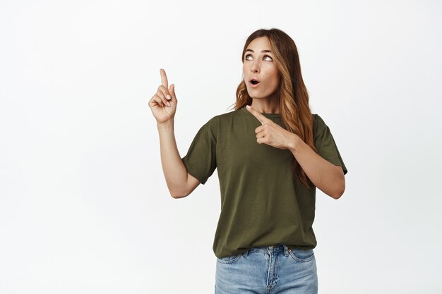 Portret van een verraste volwassen vrouw in t-shirt, wijzende vingers en kijkend naar de linkerbovenhoek