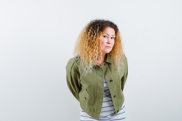 Portret van een volwassen vrouw die haar handen achter haar houdt in een groen jasje, een t-shirt en een verbaasd vooraanzicht kijkt