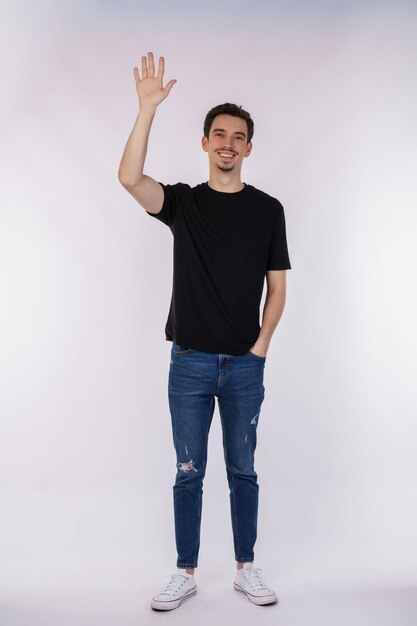 Portret van een vriendelijke, vrolijke man in een zwart t-shirt en een spijkerbroek die hallo welkome persoon zegt als een zwaaiende hand