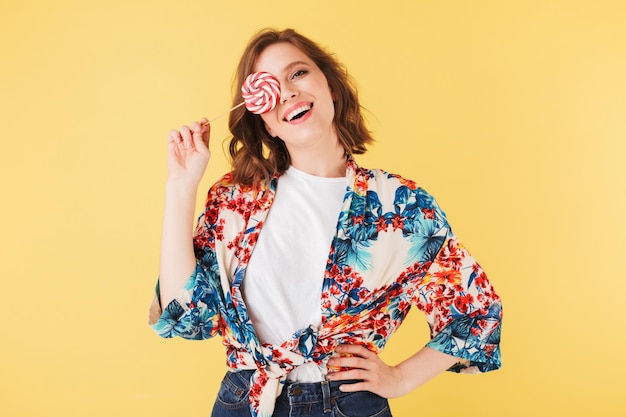 Portret van een vrij vrolijke dame in een kleurrijk shirt die staat en haar gezicht bedekt met lollysuikergoed terwijl ze vrolijk in de camera kijkt over een roze achtergrond