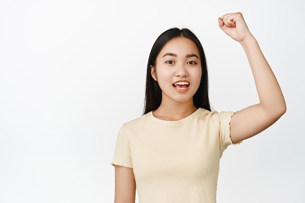 Portret van een vrolijk Aziatisch meisje dat haar hand opsteekt en protesteert en zingt en er aangemoedigd uitziet terwijl ze op een witte achtergrond staat