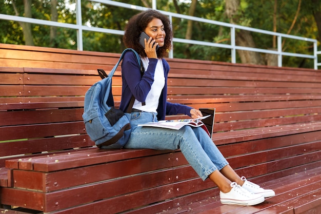 Portret van een vrolijk jong afrikaans meisje met rugzak praten op mobiele telefoon tijdens het rusten in het park, tijdschrift lezen