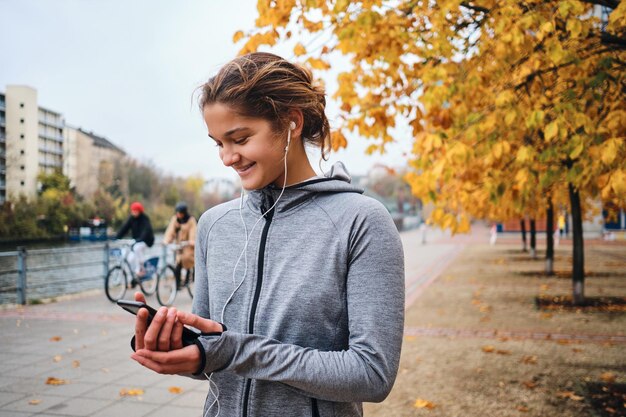 Portret van een vrolijk, sportief meisje in oortelefoons die graag een mobiele telefoon gebruikt die zich voorbereidt op hardlopen in het stadspark