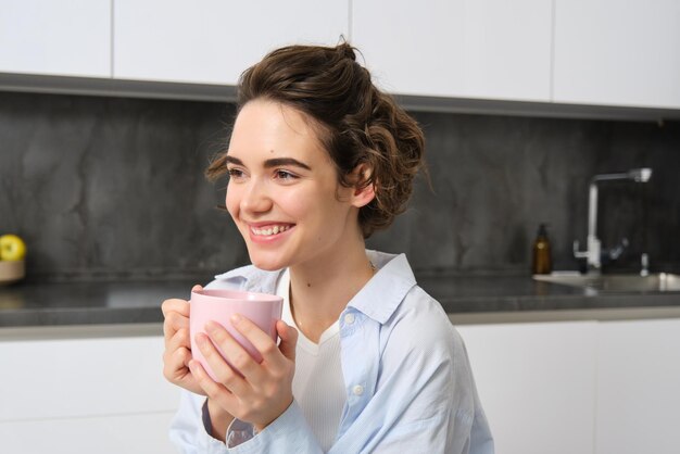 Portret van een vrolijke jonge vrouw die thuis van een kop koffie geniet, een glimlachend mooi meisje dat warm drinkt.