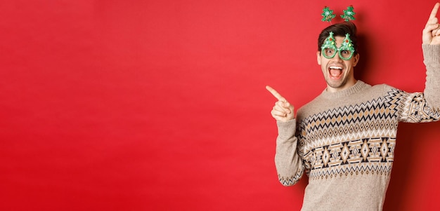 Portret van een vrolijke knappe jongen in een feestbril en een kersttrui die danst en met de vingers wijst...